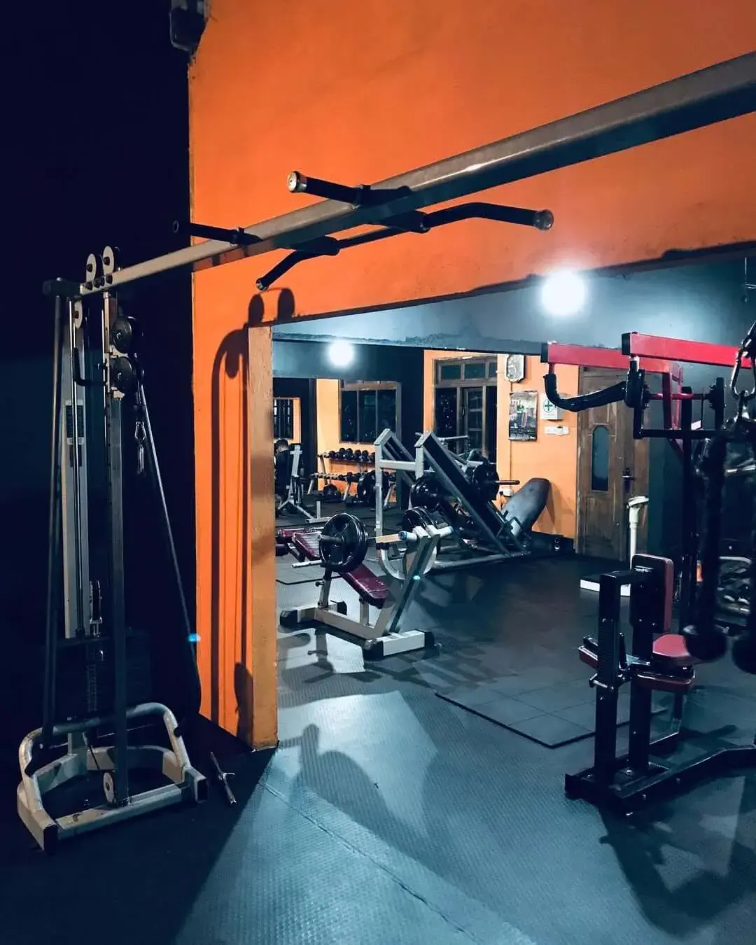 The gym floor with orange walls, a cable crossover station and red-and-black strength machines under spotlights