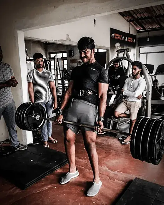 A member deadlifts a heavily loaded bar while other members watch and cheer inside the gym
